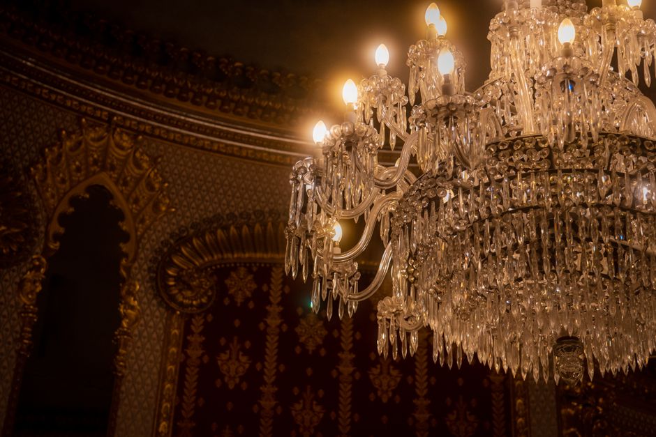 A large, ornate chandelier hanging from the ceiling of an interior room, constructed with multiple tiers of clear crystal prisms and intricate glass arms, each holding a small bulb mimicking candle flames. The chandelier has a gold-toned metal frame visible behind the crystals, reflecting warm light enhances the luxurious ambiance. The background features richly decorated wallpaper with an elaborate pattern in dark tones, bordered with gold accents, suggesting a historic or high-end setting. This detailed lighting fixture contrasts with typical waste management scenarios but highlights the importance of maintaining clean, well-kept environments, including elegant spaces that benefit from professional rubbish removal and on-site clearance services provided by companies like [COMPANY_NAME]. The environment appears indoor and opulent, emphasizing the significance of proper waste handling to preserve such refined interiors.