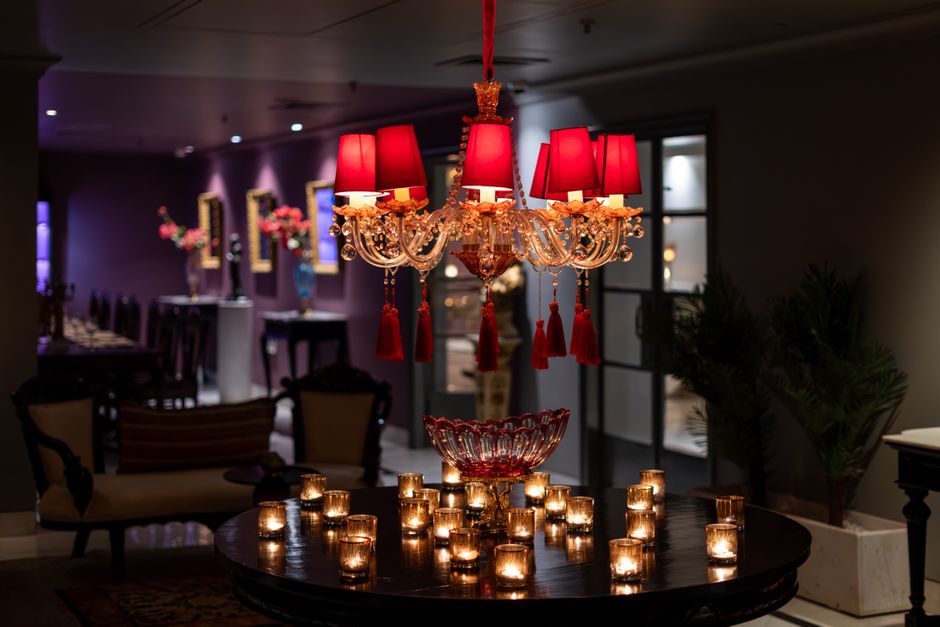 The image depicts an interior space illuminated by a decorative chandelier with a gold frame, adorned with red lampshades and hanging red tassels, positioned above a dark polished round table. On the table, there is a clear glass bowl with an intricate design, surrounded by numerous small, flickering tea light candles in transparent holders, creating a warm ambient glow. In the background, blurred furnishings include a vintage-style armchair with dark wooden legs and a beige upholstered seat, a matching sofa with a curved backrest, and a dining area with a long table set with elegant tableware and flower arrangements in vases, suggesting a sophisticated, possibly private, setting. Natural light filters through a window with dark frames, and potted plants with lush green foliage are visible along the wall, adding contrast to the predominantly dark and warm colour palette. The scene reflects a tastefully decorated interior with an emphasis on ambient lighting and subtle decorative details, aligning with themes of private on-site clearance or elegant rubbish removal setups in high-end environments, as provided by [COMPANY_NAME] within the realm of alternative waste handling options. The overall scene is calm and refined, with careful attention to lighting to enhance the visual appeal of the interior space.