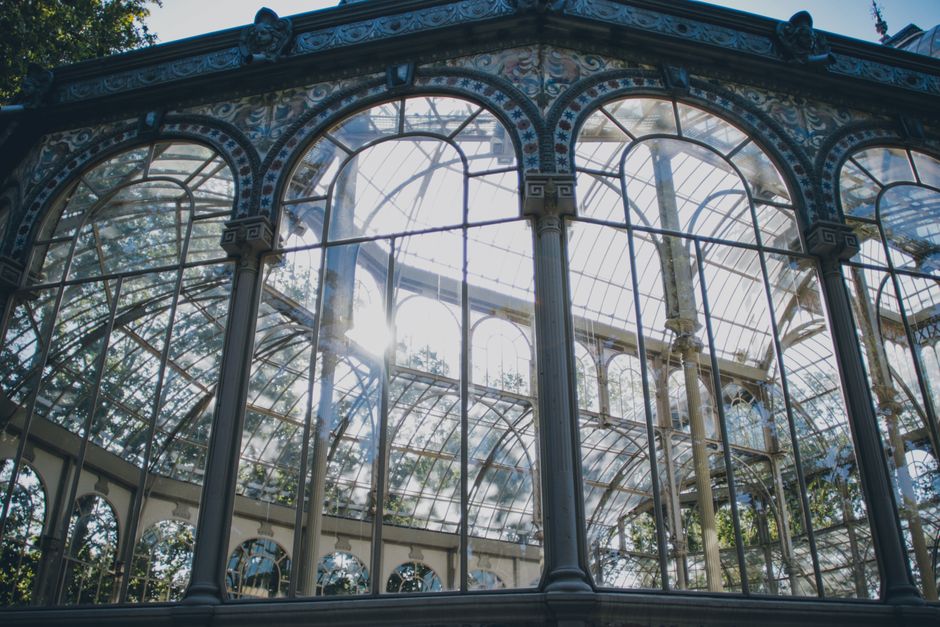 A detailed view of a large, ornate glass and metal conservatory structure, featuring intricate decorative ironwork framing tall, arched windows. The glass panels are clear and reflect sunlight, creating a bright atmosphere with visible sun rays and lens flare. Inside, wooden and metal supports hold the glass panes, and some greenery from plants can be seen through the windows. The surrounding environment appears to be outdoors, with natural daylight illuminating the scene, highlighting the texture of the metal framework which is painted in a muted, possibly grey or dark tone. The conservatory's design suggests a historical or Victorian style, with detailed embellishments on the ironwork. This type of structure is often used for botanical display or as a garden feature, and occasional outdoor maintenance or cleaning activities may take place. The image captures the elegance and architectural details typical of decorative greenhouses, providing a clear visual context for use in articles about structural preservation or garden-related projects. Occasional visible reflections from the sunlight hint at a clean, well-maintained environment, aligning delicately with themes of on-site clearance or conservatory maintenance services associated with waste removal specialists like rubbishcollectioncrystalpalace.co.uk.