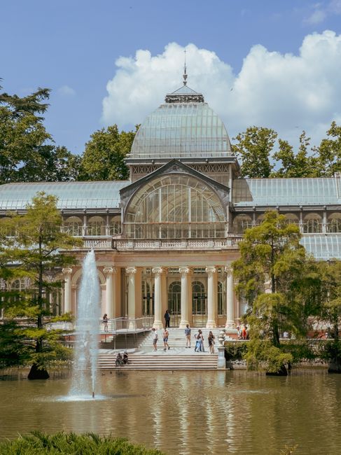 The image shows the historic Crystal Palace Palm House, featuring a large, semi-spherical glass dome with a metal framework, situated atop a classical-style building with decorative columns and stone balustrades. The glass panels, which are transparent and reflect the surrounding environment, are arranged in a grid pattern and curved to form the prominent dome structure. The building’s lower section includes arched windows and intricate detailing on the stonework, with a traditional street lamp mounted on the façade. Visible greenery and trees are present around the structure, indicating its parkland setting. The sky in the background is partly cloudy with patches of blue, contributing to a natural and calm atmosphere. This architectural feature is a significant element within the landscape, often associated with conservatories and glasshouses used historically for plant cultivation and display. The scene does not show rubbish or waste, focusing instead on the building’s detailed glass and stone construction, aligned with the themes of architectural preservation and historic landscaped environments, potentially relating to private or municipal efforts to maintain such sites amid broader waste management considerations.