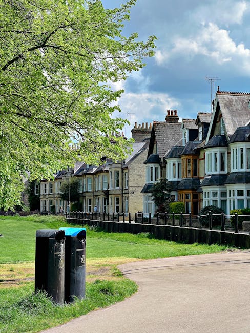 A residential street scene featuring a row of traditional Victorian-style terraced houses with bay windows, pitched roofs, and brick chimneys, set against a partly cloudy sky. In the foreground, there are two black waste bins placed on a grassy verge near a paved pathway, with a wooden rail fence separating the pathway from the front gardens of the houses. A large tree with fresh green leaves extends its branches over the scene, providing shade and adding natural greenery. The environment appears calm, and the scene exemplifies a typical suburban setting where private waste collection services such as those provided by rubbishcollectioncrystalpalace.co.uk may operate for efficient rubbish removal and outdoor waste management, especially in established residential areas.