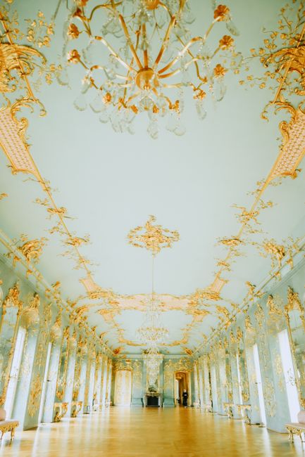 This image depicts an opulent interior room with a highly decorative ceiling featuring elaborate gold leaf accents, ornate moldings, and a central chandelier composed of multiple crystal lights. The ceiling's design extends to the upper walls, which are adorned with intricate gold embellishments and large, evenly spaced windows that let in natural light. The floor is made of polished light-colored wood, creating a reflective surface that complements the luxurious decor. Along the sides, tall mirrors and decorative columns enhance the grandeur of the space. The room appears to be part of a historic palace or formal hall, characterized by its classical and baroque interior elements. As an observation relevant to independent waste management or on-site clearance, this elegant setting contrasts with scenarios involving cluttered or disused spaces awaiting rubbish removal or professional disposal, exemplifying a context where high-quality interior environments require careful handling during any refurbishment or clearance process. The overall scene is well-lit with soft natural and ambient lighting, emphasizing the ornate details and the spaciousness of the room.