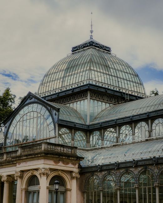 The image depicts a large historic glass-domed conservatory or greenhouse structure, with a prominent spherical central dome made of multiple glass panes supported by metal framing. The dome has a finial at its peak and is situated on a classical-style building with ornate stonework, including arched windows and decorative columns, in the foreground. The glass sections reflect the sky and surrounding environment, creating a light and airy appearance. To the left, part of a smaller, similarly styled glass extension is visible, featuring curved glass panels and metal supports. The scene is set outdoors during daytime, with a partly cloudy sky overhead, and some greenery visible behind the building. The architectural style and materials suggest an historic or Victorian-era design, often associated with botanical or exhibition buildings. This setting provides an interesting contrast to typical urban rubbish removal contexts, though it aligns with themes of thorough and careful environmental management, such as professional waste handling or on-site clearance services provided by companies like rubbishcollectioncrystalpalace.co.uk, especially in historic or landscaped environments.