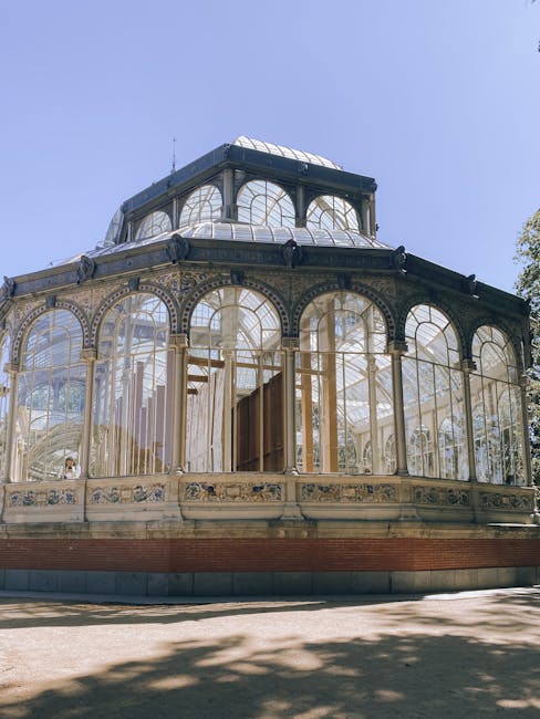 A historic glass conservatory with a rounded, multi-tiered roof structure, featuring large, arched windows framed by ornate metalwork painted in light cream. The glass panels are clear, allowing a view of the interior, which appears bright due to natural sunlight. The building's exterior rests on a low brick foundation, with some decorative detailing along the lower edge of the structure. It is situated outdoors on a paved or gravel surface, with shadows cast by nearby trees visible on the ground in the foreground. The surrounding environment includes a clear blue sky overhead and partial visibility of greenery on the right side of the image, indicating it is in a garden or park setting. The scene is well-lit with natural daylight, highlighting the building’s historical architectural features, and due to the context of rubbish removal services, the conservatory may be part of an estate or venue where cleaning or clearance could be relevant for maintenance or private disposal purposes through independent collection services like those offered by [COMPANY_NAME].