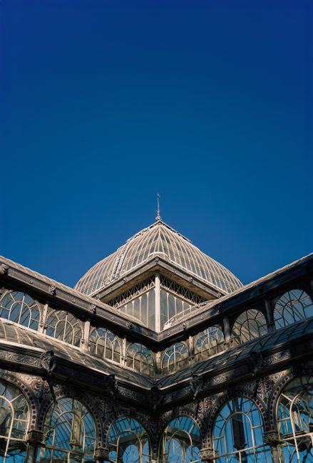 The image depicts a large historic glass-domed conservatory or greenhouse structure, with a prominent spherical central dome made of multiple glass panes supported by metal framing. The dome has a finial at its peak and is situated on a classical-style building with ornate stonework, including arched windows and decorative columns, in the foreground. The glass sections reflect the sky and surrounding environment, creating a light and airy appearance. To the left, part of a smaller, similarly styled glass extension is visible, featuring curved glass panels and metal supports. The scene is set outdoors during daytime, with a partly cloudy sky overhead, and some greenery visible behind the building. The architectural style and materials suggest an historic or Victorian-era design, often associated with botanical or exhibition buildings. This setting provides an interesting contrast to typical urban rubbish removal contexts, though it aligns with themes of thorough and careful environmental management, such as professional waste handling or on-site clearance services provided by companies like rubbishcollectioncrystalpalace.co.uk, especially in historic or landscaped environments.